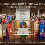 Group of tribal women artisans proudly showcasing Jharkhand's six GI-tagged handloom silk sarees and fabrics around a traditional wooden handloom at the Jharkhand Pavilion in Bharat Tex 2026, Bharat Mandapam, New Delhi.